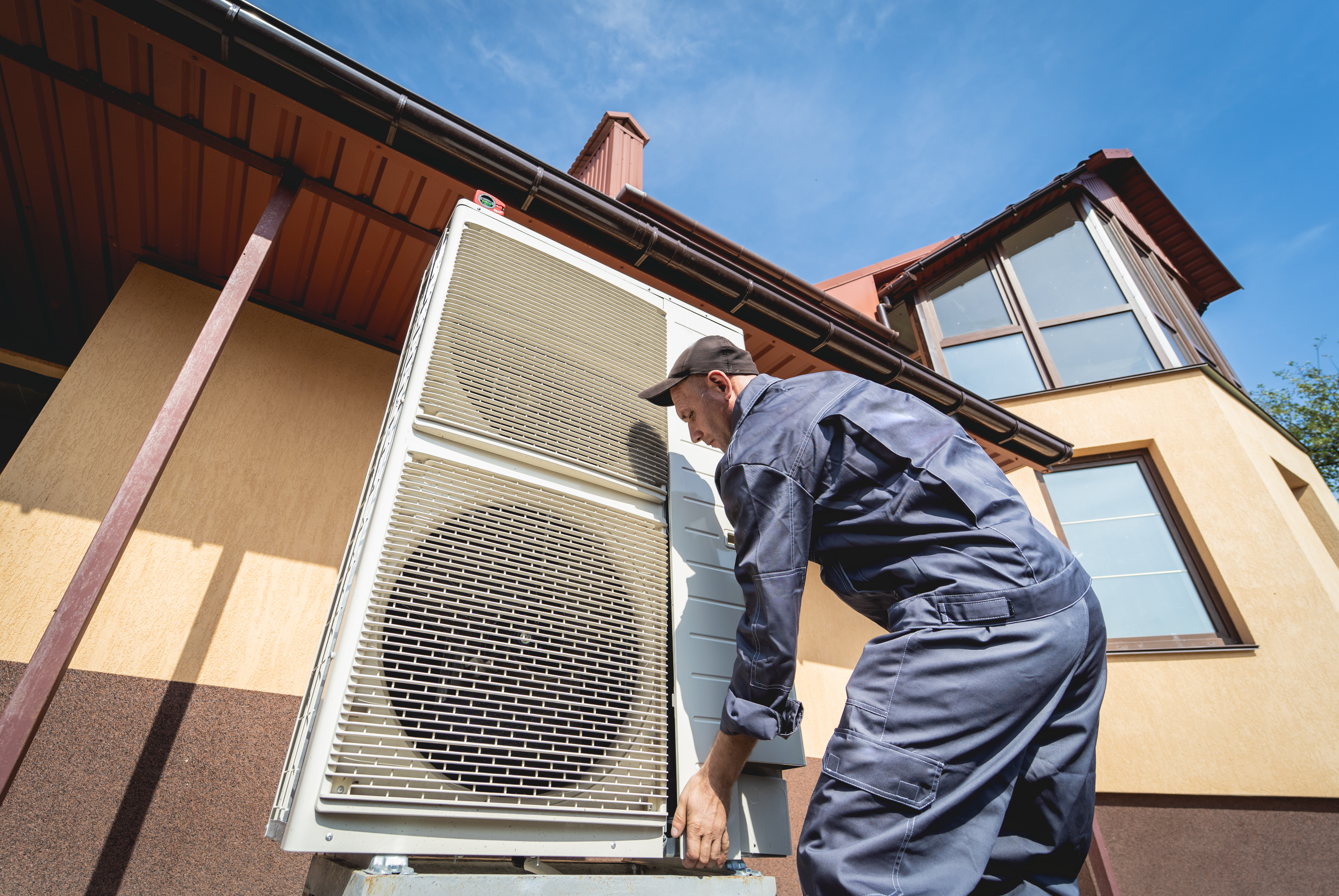 A ház mellett férfi szereli be a hőszivattyú kültéri elemét.