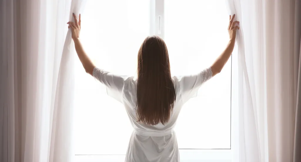 Nő az ablaknél háttal áll és húzza szét két karjával a függönyt